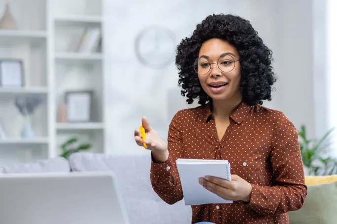 Curly-haired woman takes notes while engaging in virtual meeting.