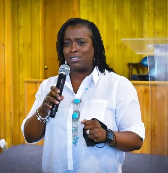 Woman with microphone presents indoors wearing white and turquoise.