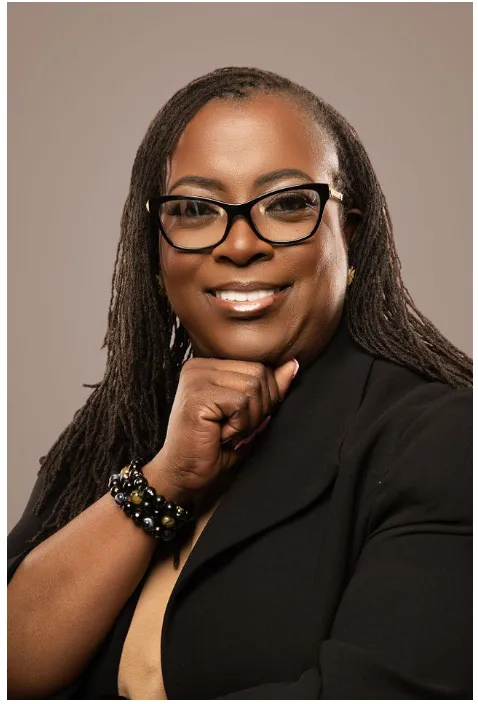 Confident woman in a business suit with glasses, smiling and resting her chin on her hand. Professional portrait suggesting approachability and expertise.